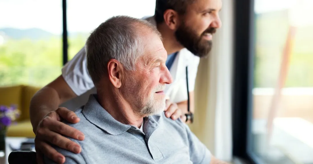 Person sitting in a chair looking out a window with a healthcare provider next to them with their hands on the person's shoulders Person sitting in a chair looking out a window with a healthcare provider next to them with their hands on the person's shoulders